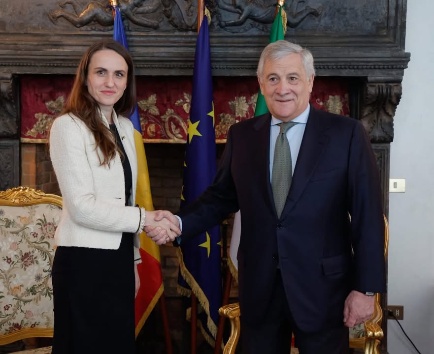 Oana Țoiu, ministra de Externe a României, dă mâna cu un oficial italian în fața drapelelor României, Italiei și Uniunii Europene